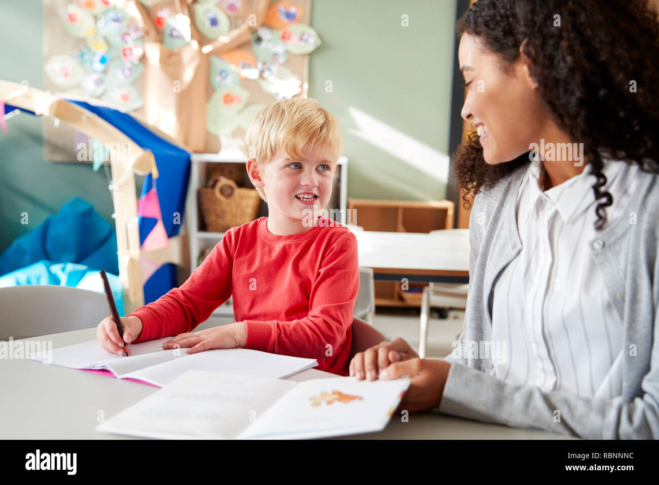 Female infant school teacher working one on one with a young white schoolboy, sitting at a table in a classroom writing, looking at each other, close up Stock Photo