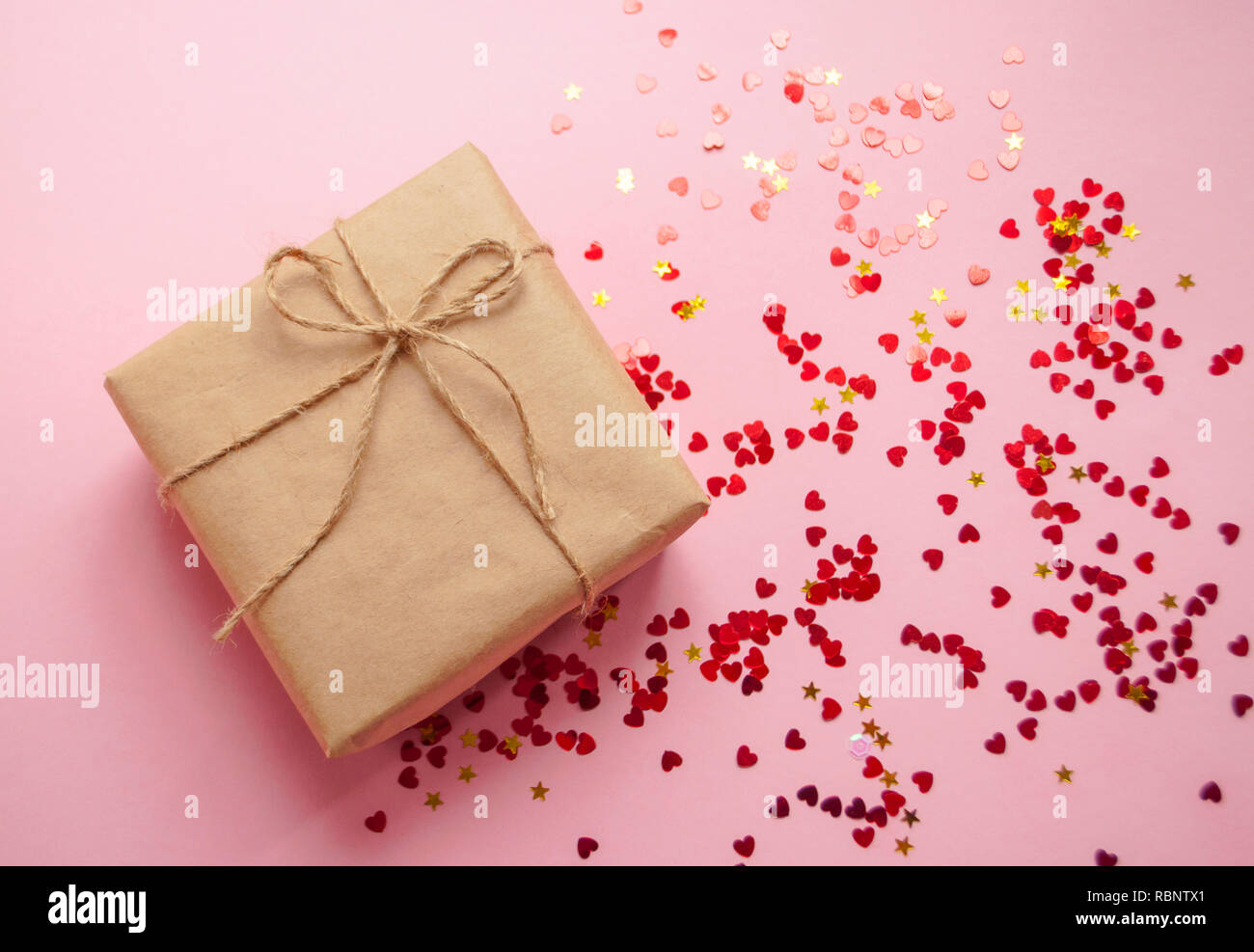 Gift box wrapped in brown colored craft paper and tied with rope on pink background with heart shape red confetti. Stock Photo