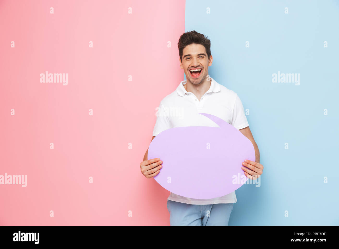 Excited young man standing isolated over two colored background, holding empty speech bubble Stock Photo
