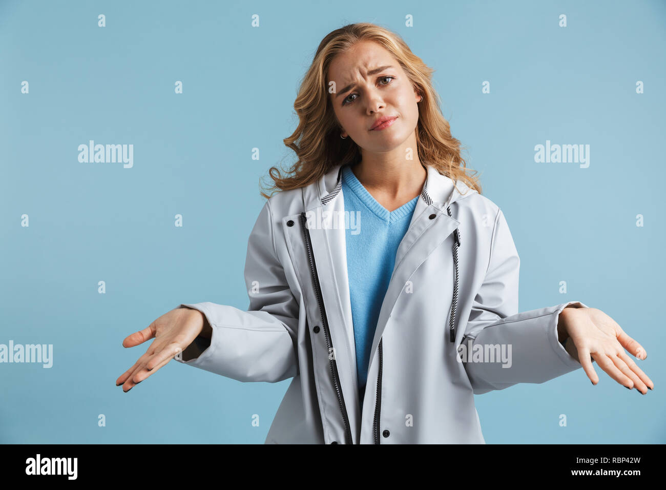 Confused young girl wearing raincoat standing isolated over blue background, shrugging shoulders Stock Photo
