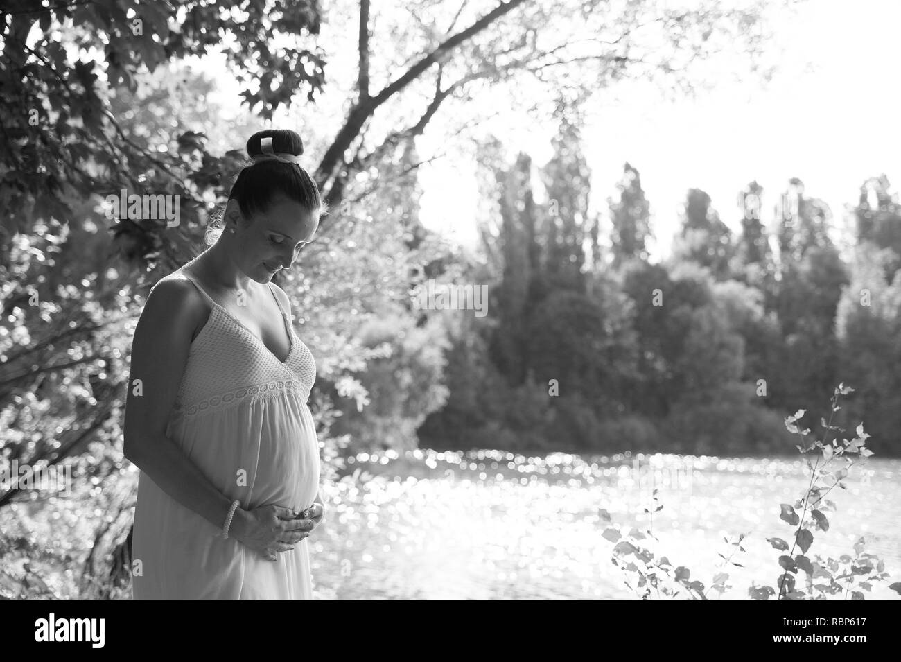 Milan, Italy - 20 july 2018: black and white portrait of young expecting woman with white dress holding her belly outdoors with sun reflecting on lake Stock Photo