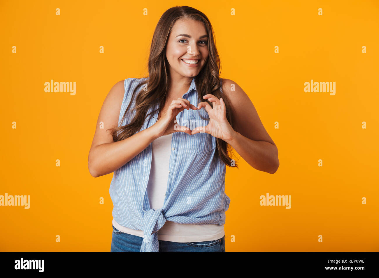 Smiling young overweight woman standing isolated over yellow background, showing heart gesture Stock Photo