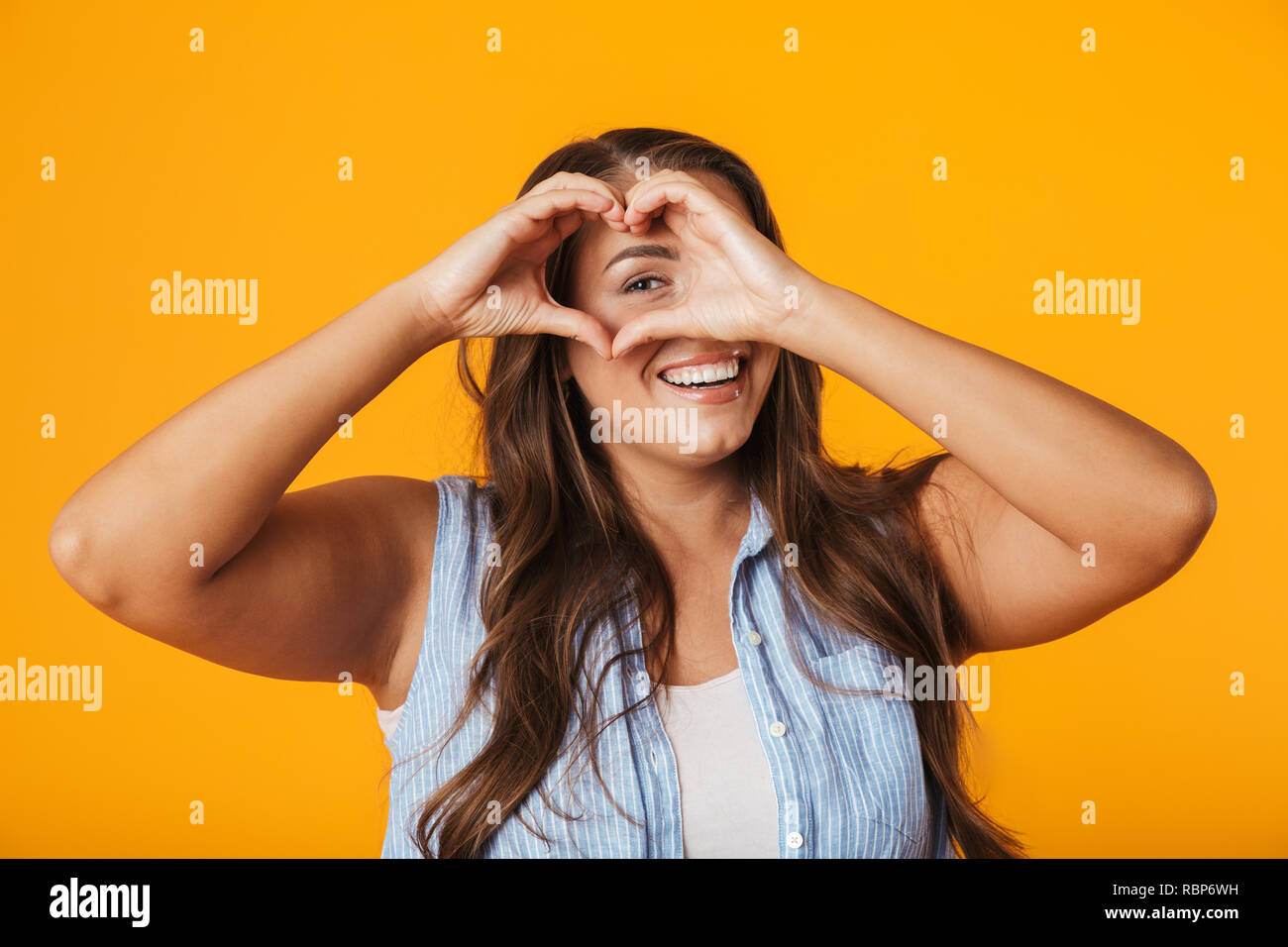 Smiling young overweight woman standing isolated over yellow background, showing heart gesture Stock Photo