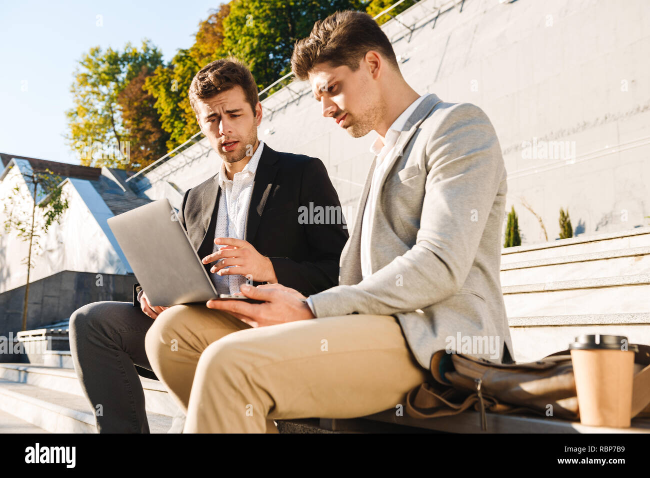 Two confident businessmen using laptop computer while sitting on a bench outdoors Stock Photo