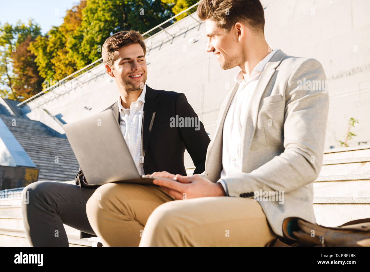Two confident businessmen using laptop computer while sitting on a bench outdoors Stock Photo