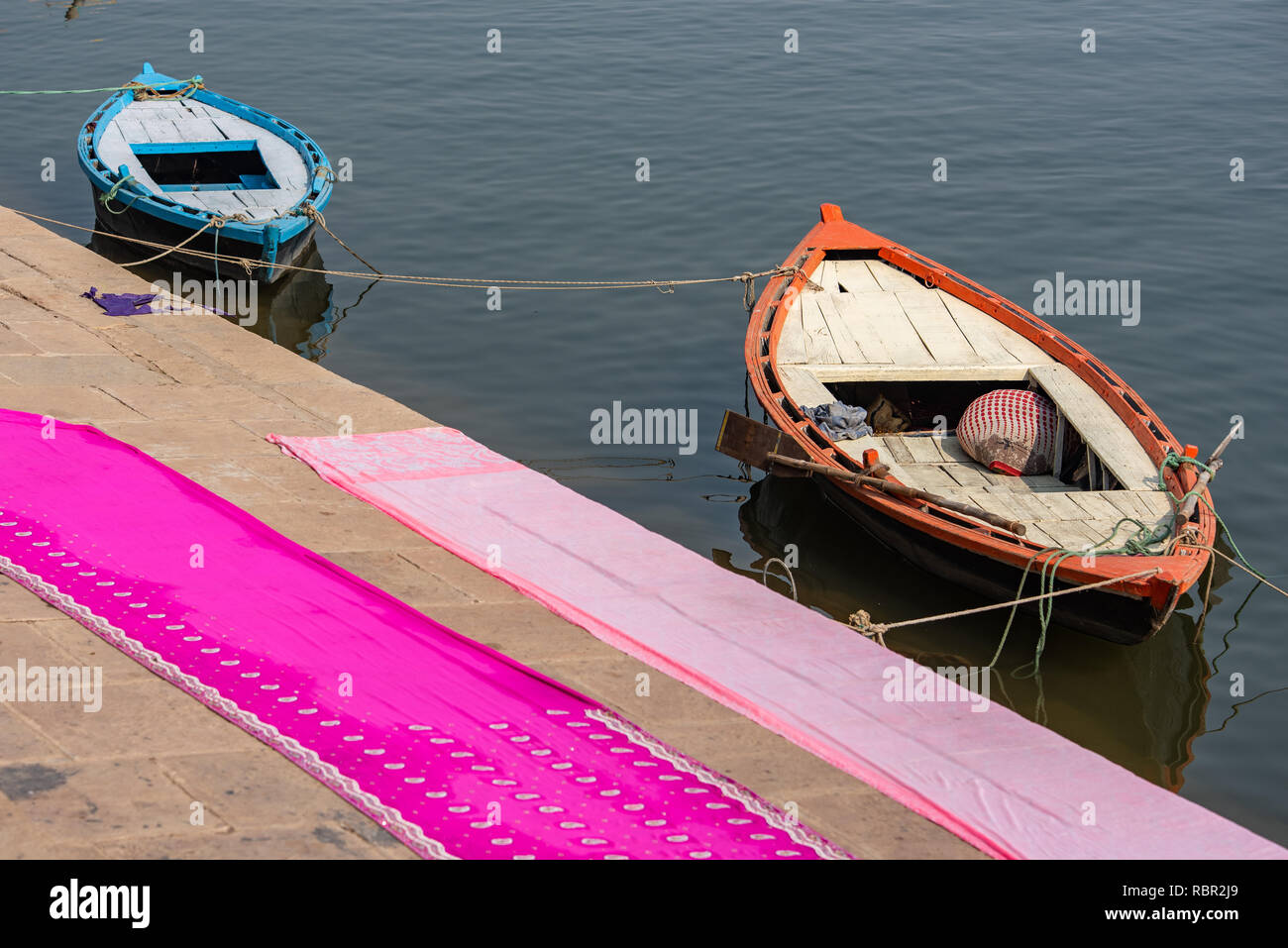 Two boats on the Ganges in the sunshine with two long dhotis or sarees drying on the paving in the foreground: photo taken in Varanasi, India Stock Photo