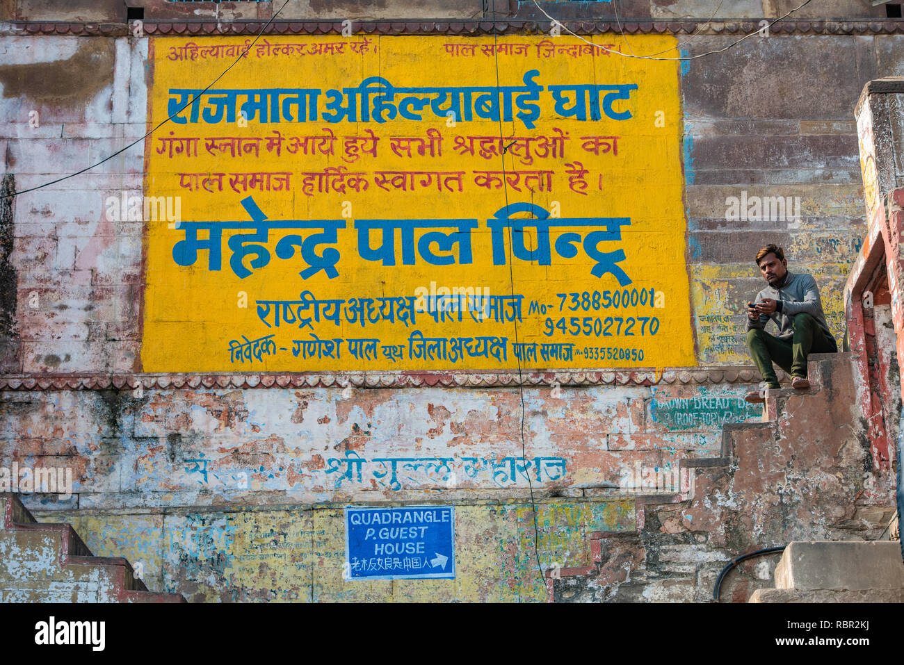 A sign on the wall giving details about a local political party 'Pal Samaj' in Varanasi, India with a man sitting on a step next to it. Stock Photo