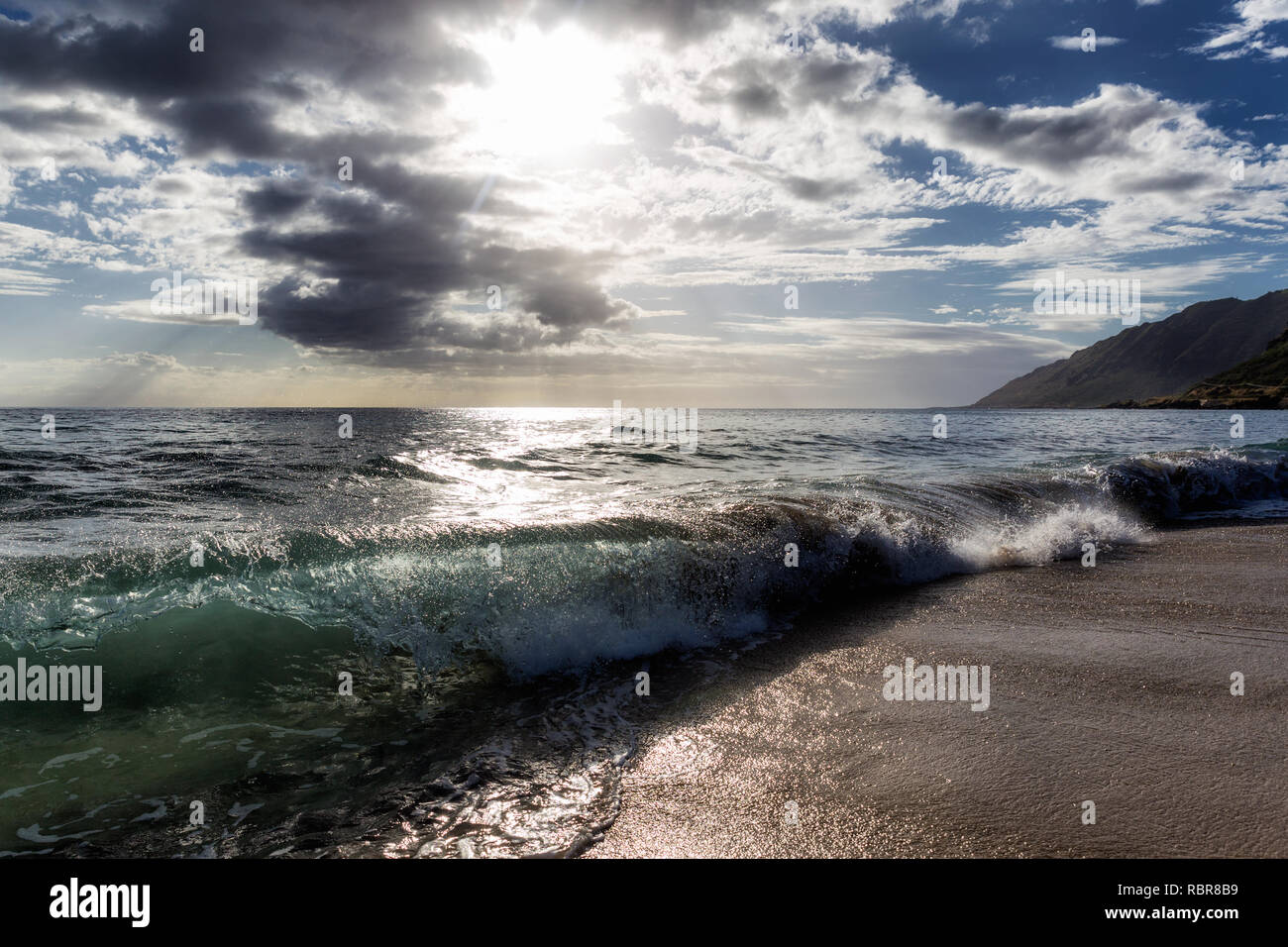 Sun shining through the clouds at sandy beach with waves Stock Photo