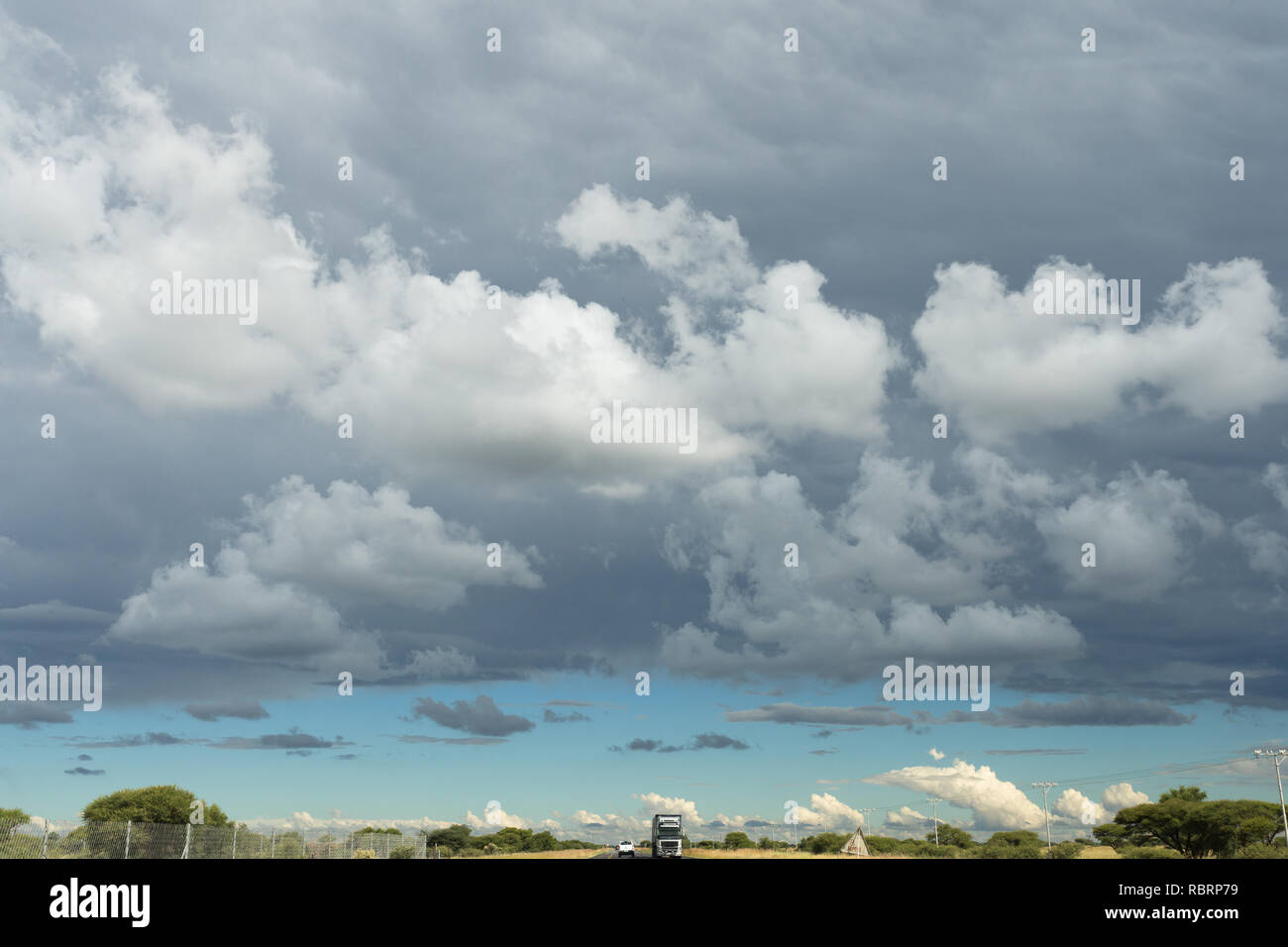 Cumulus or puffy clouds in the sky and below a long road stretching into infinity with vehicles in the North West Province of South Africa Stock Photo