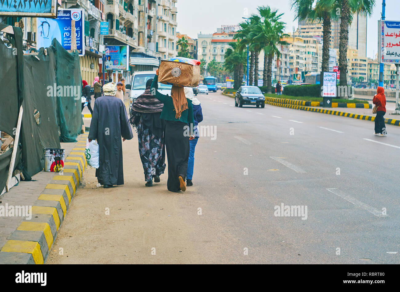 ALEXANDRIA, EGYPT - DECEMBER 19, 2017: The group of locals walks along the road in Corniche Avenue, young woman carries the bag on her head, on Decemb Stock Photo