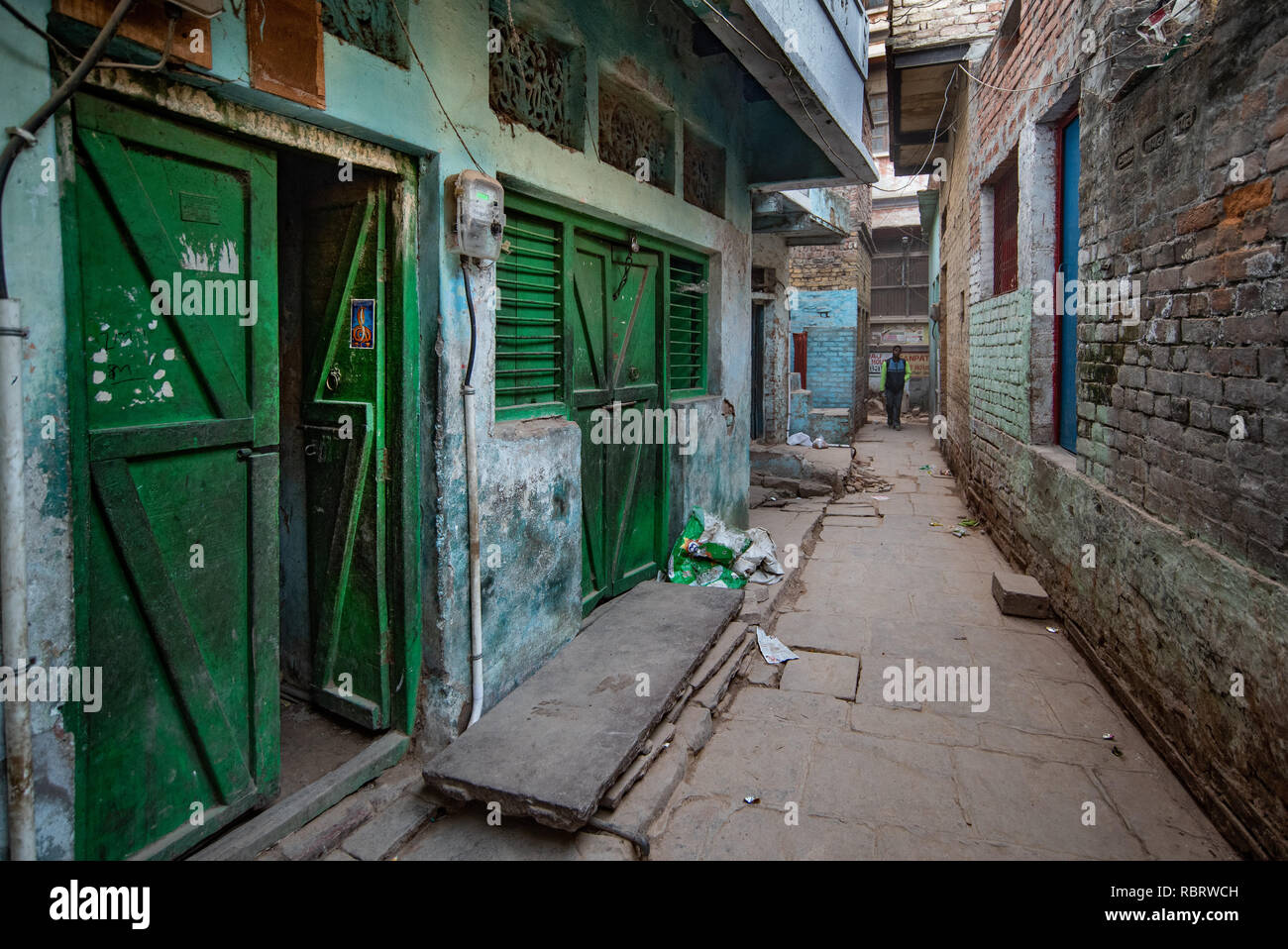A narrow and organic street in Varanasi, India with green coloured doors to one side and a brick wall opposite. Stock Photo