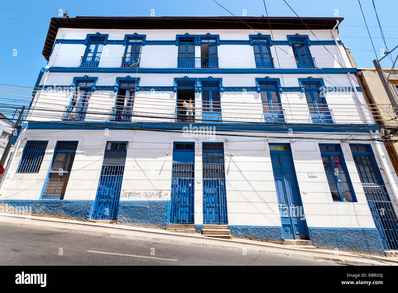 Blue and white housing block on a steep hillside location, by a street, in Valparaiso, Chile, with one of the residents reading a paper. Stock Photo