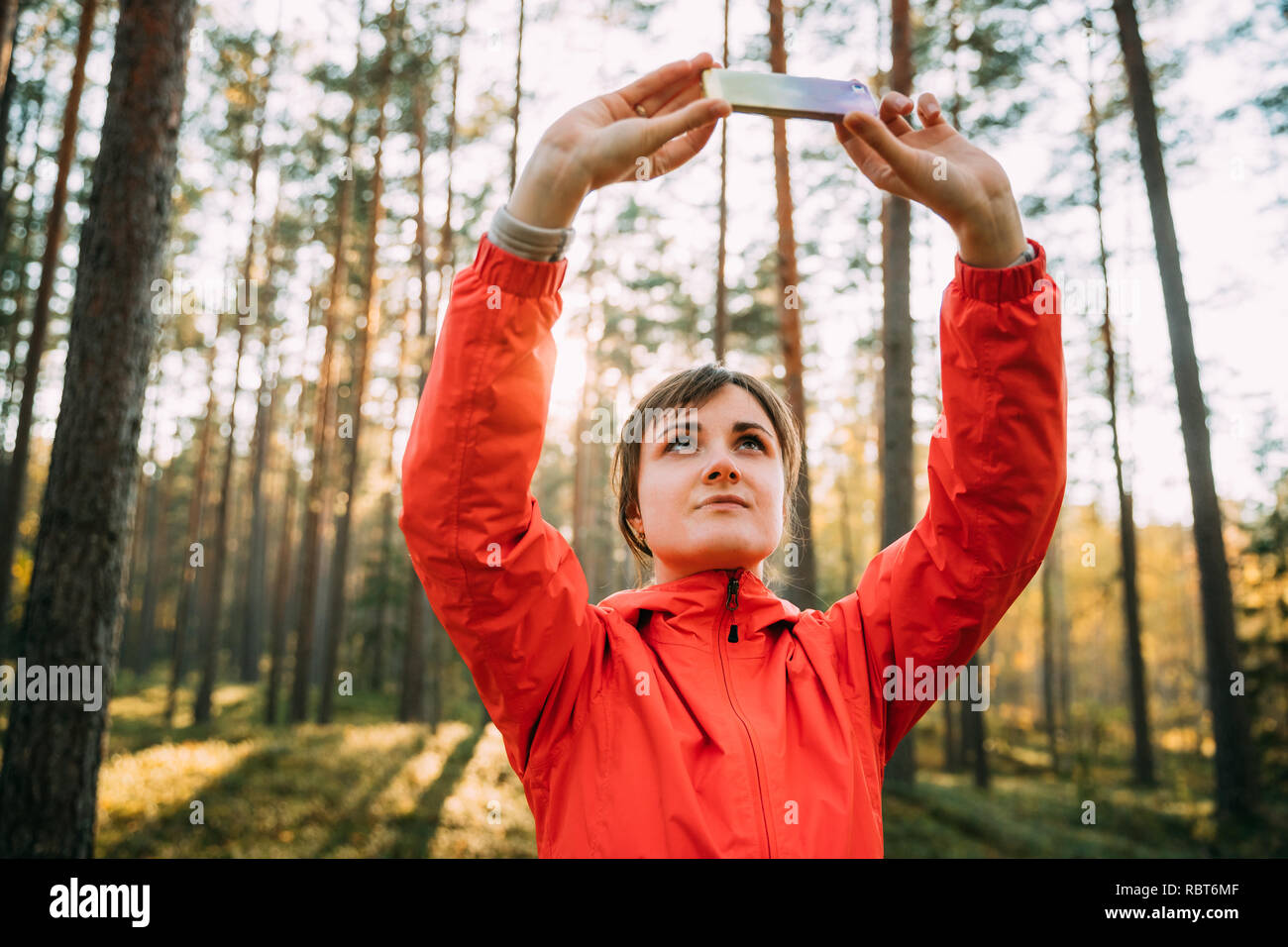 Active Young Adult Beautiful Caucasian Girl Woman Dressed In Red Jacket Taking Photos On Smartphone In Autumn Sunset Green Forest. Active Lifestyle In Stock Photo