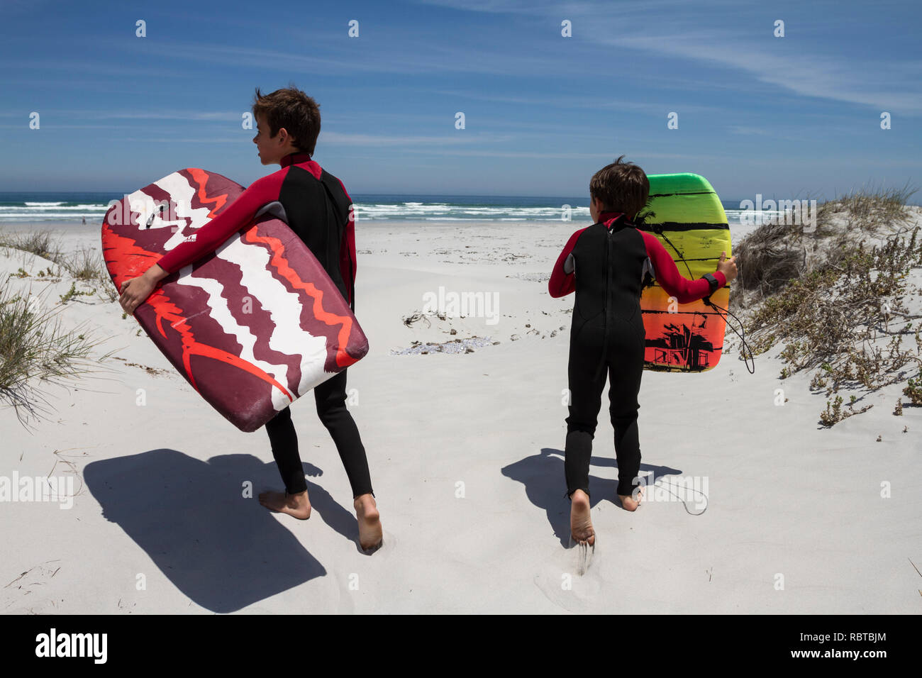 Two young boys in wet suits cross a coastal sand dune with body boards Stock Photo