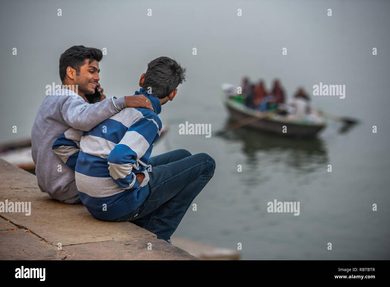 Two friends sitting on the Ghats chatting with each other while speaking on the phone and watching boats passing by on the Ganges in Varanasi, India Stock Photo