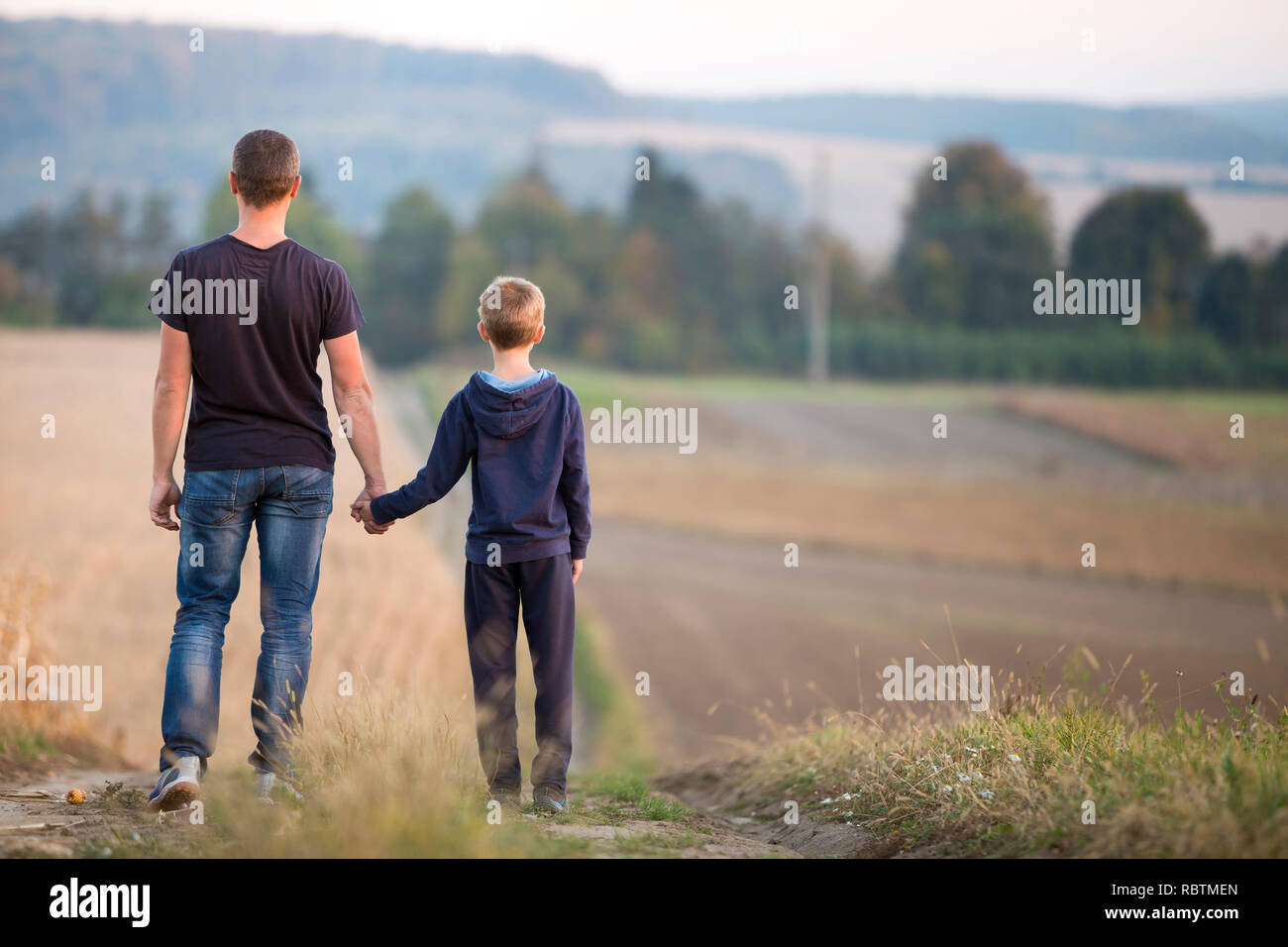 Back view of young father and son walking together holding hands by grassy field on blurred foggy green trees and blue sky background. Active lifestyl Stock Photo