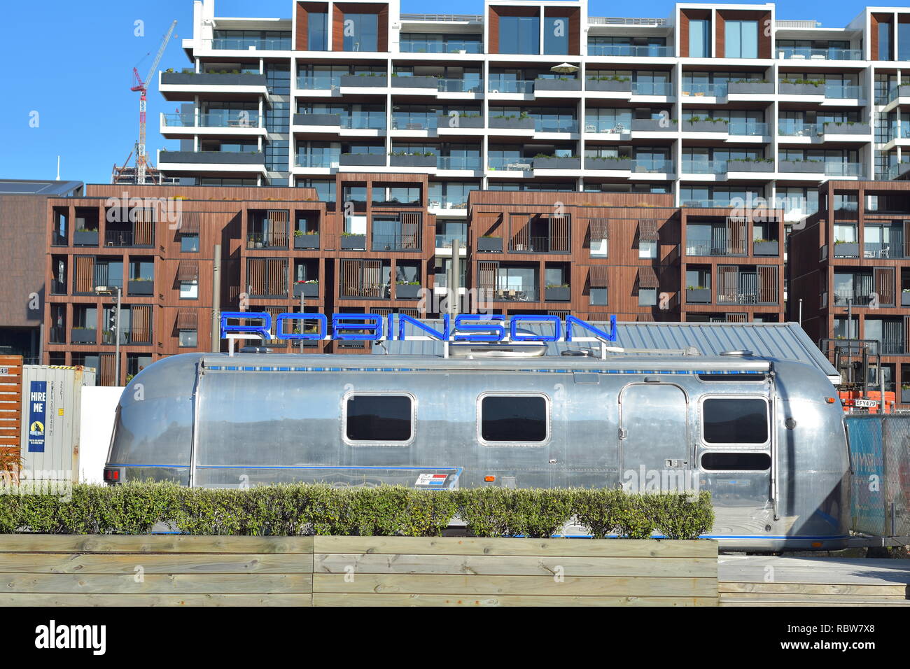 Large shiny polished aluminum caravan with big blue sign on top of it in front of apartment buildings of varying height. Stock Photo