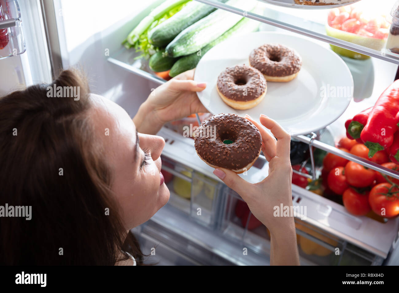 Young Happy Woman Taking Donut From Plate In Refrigerator Stock Photo