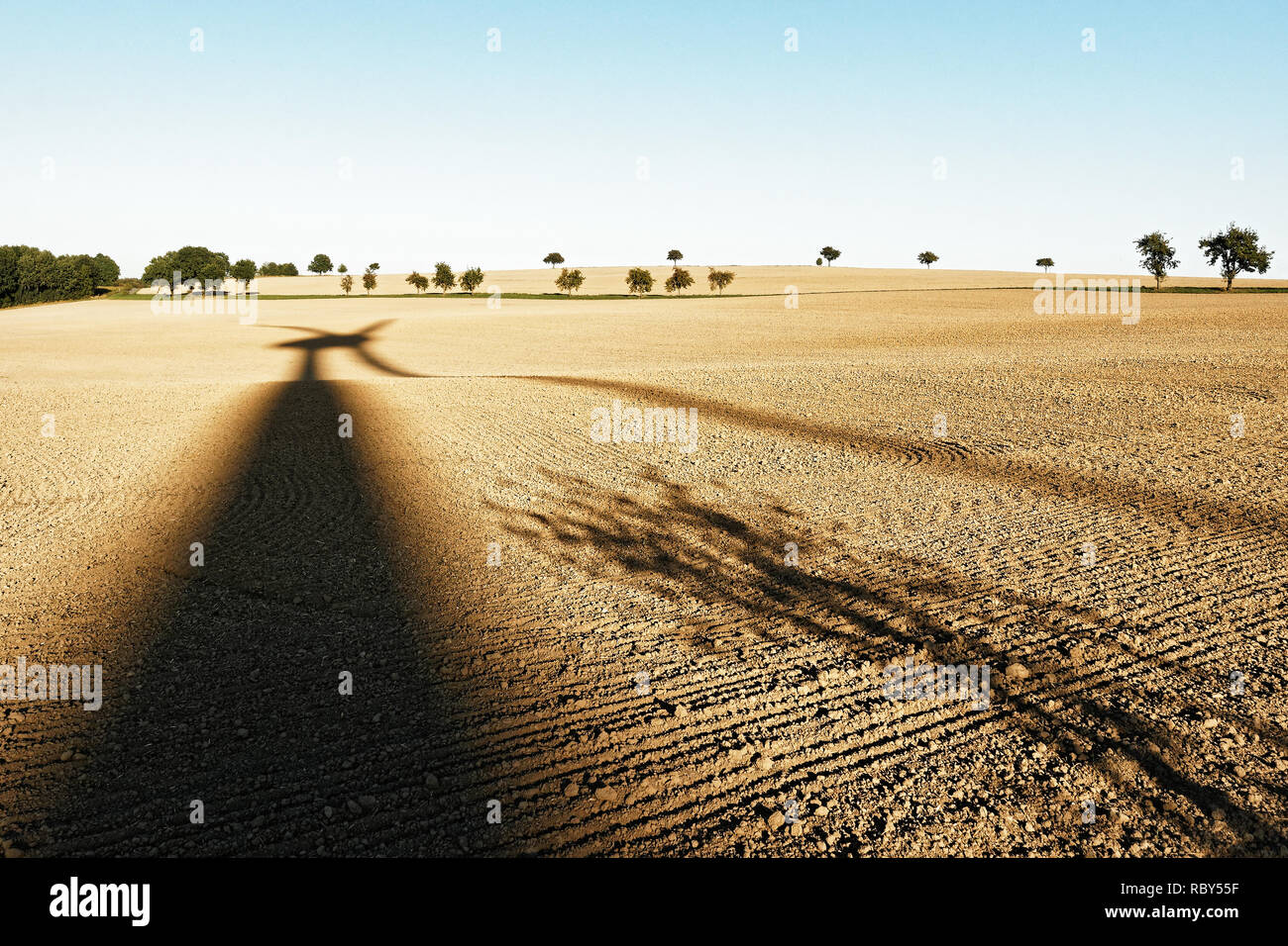 Long shadow of a windmill on a harvested field in a hilly landscape, single trees in the background, late summer mood - Location: Germany, Saxony Stock Photo
