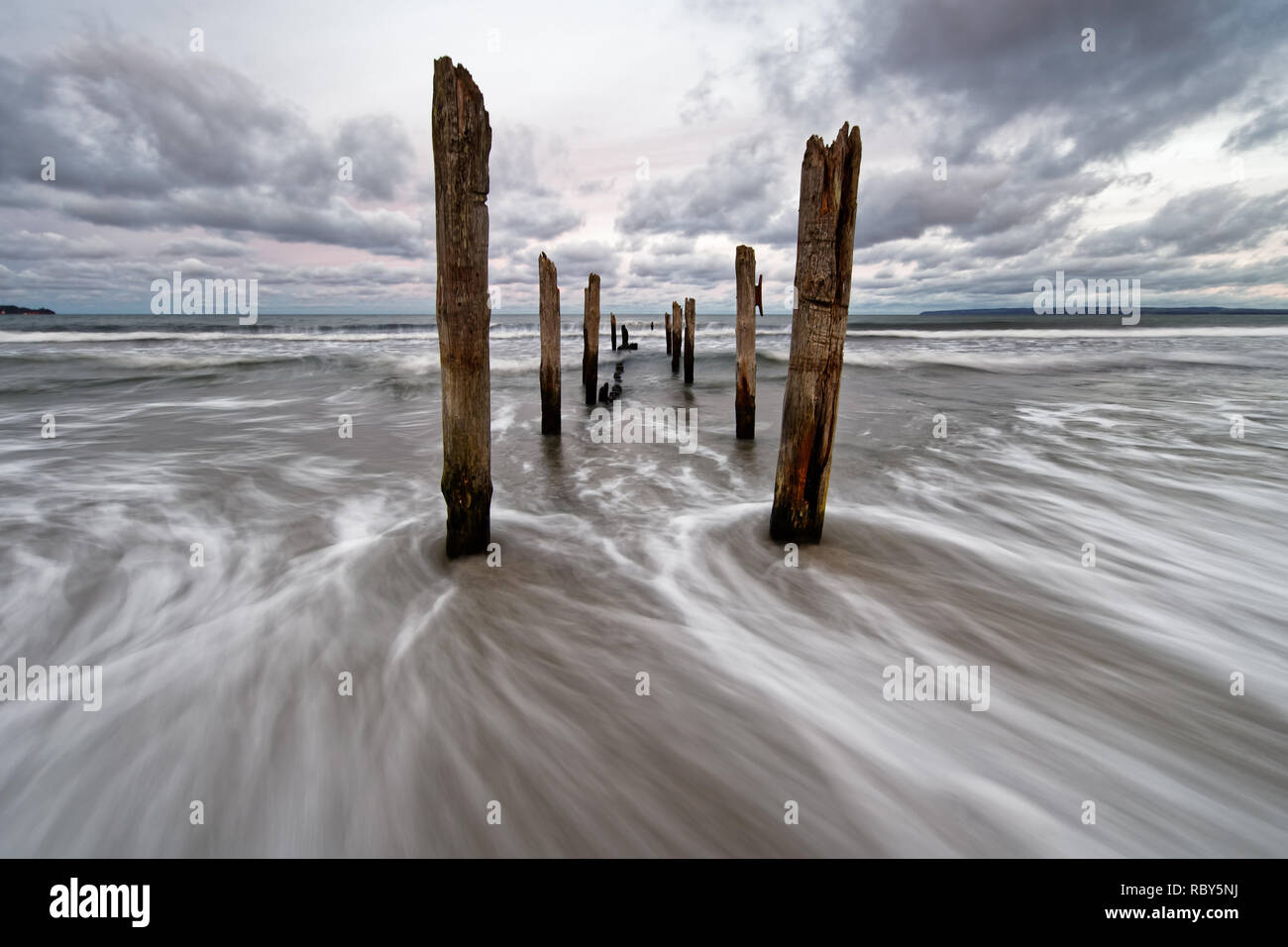 Wooden poles on Baltic Sea beach in high contrast evening light with clouds in windy weather, water movement in long exposure - Location: Baltic Sea,  Stock Photo