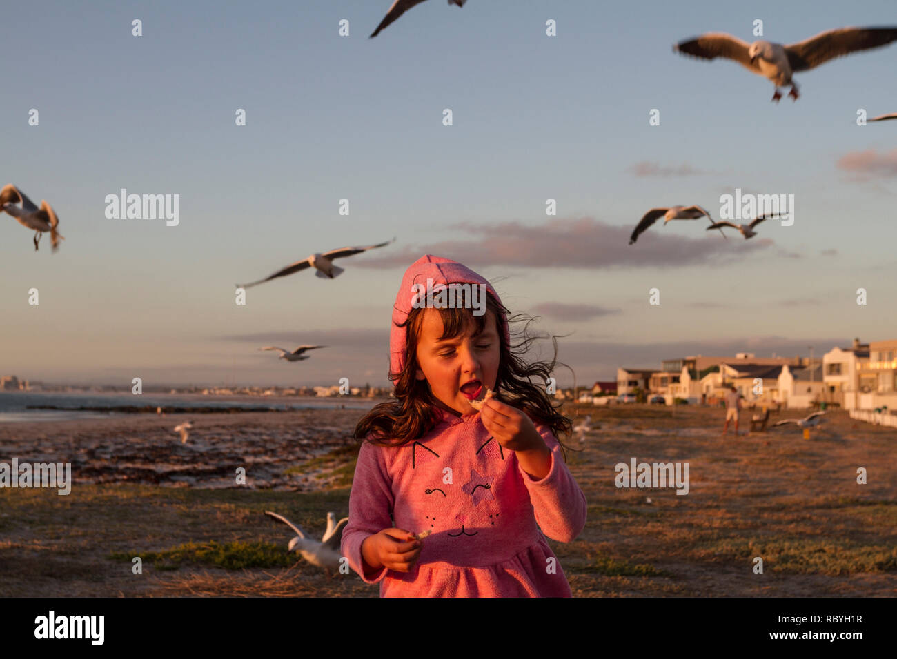 Young girl dressed in pink eating bread on the beach with seagulls flying above Stock Photo