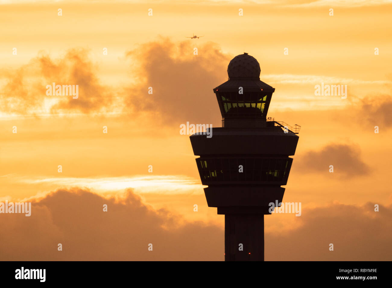 Amsterdam Schiphol International Airport control tower with a plane landing in the background during sunset. Stock Photo