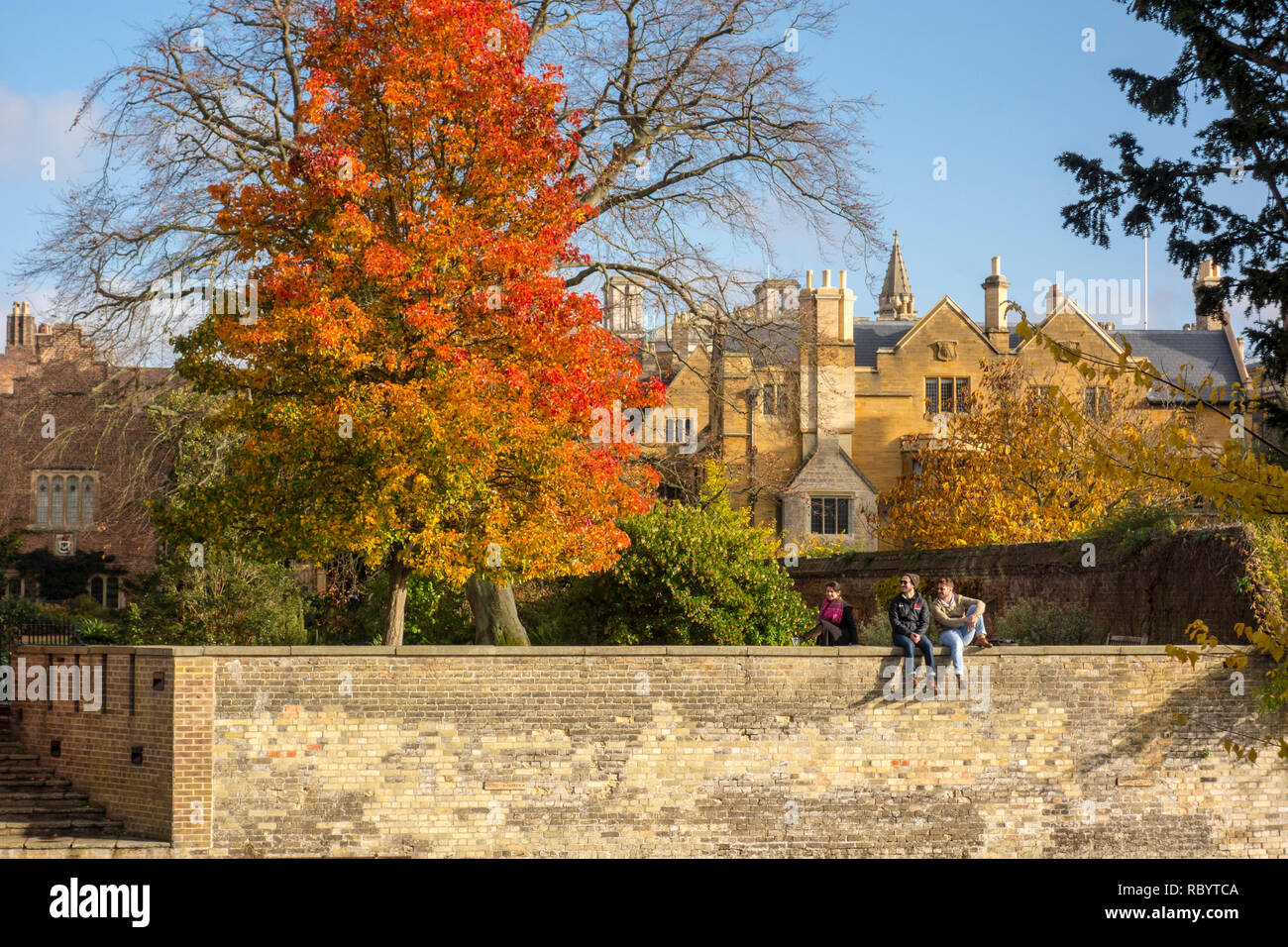 Students sat on a wall on the bank of the River Cam outside Clare College in autumn, Cambridge, UK Stock Photo