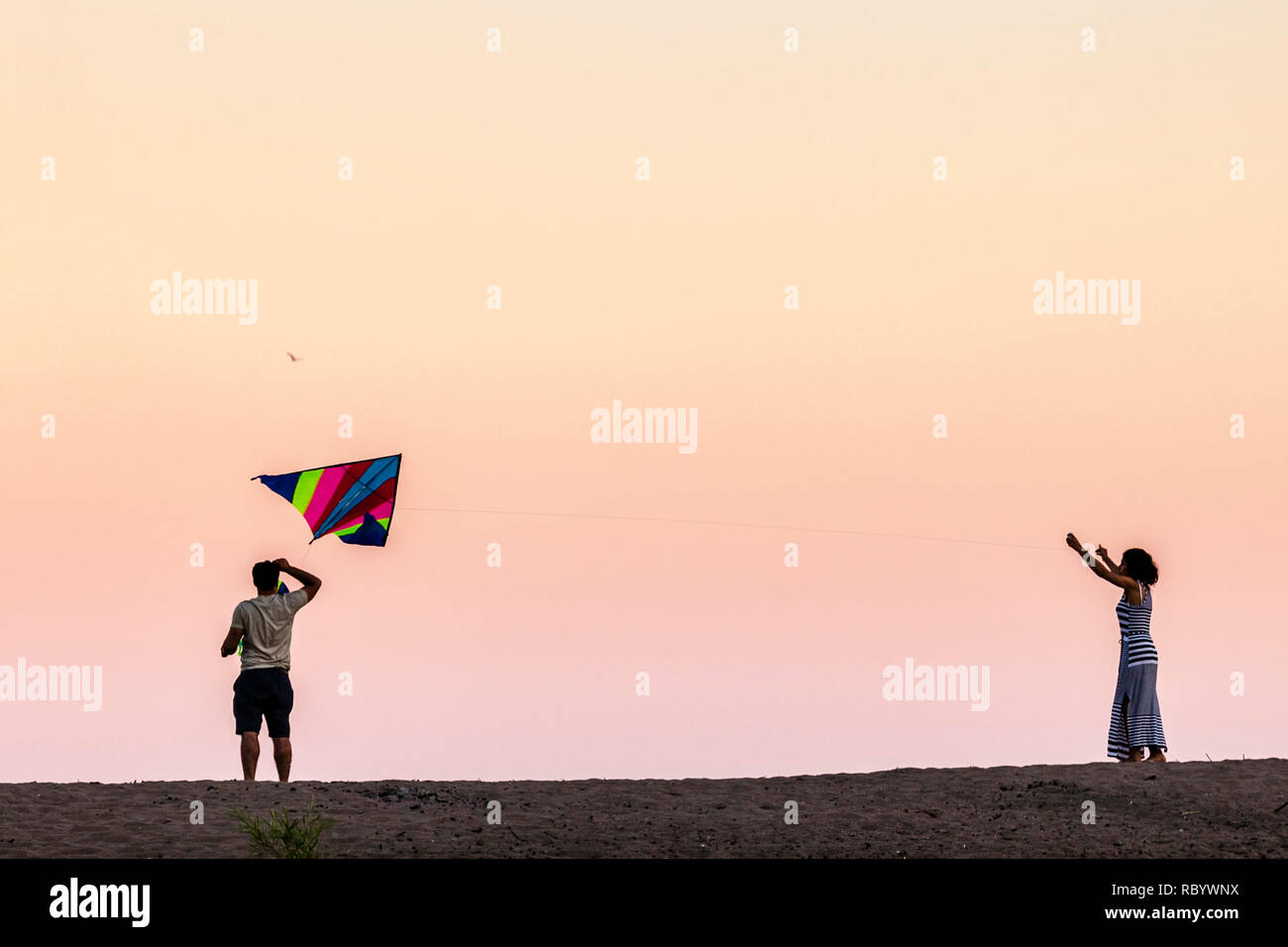 Stock Photo of A young couple playing together, flying a kite on a beach with a beautiful sunset sky. Stock Photo