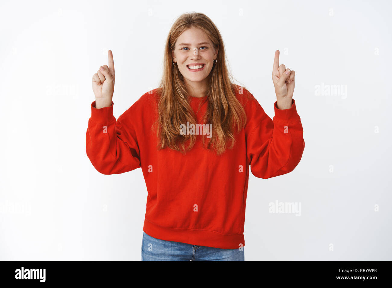 Charismatic charming optimistic young european girl with chubby cheeks, blue eyes and freckles smiling joyfully raising hands to point up at copy space posing in red stylish sweater over gray wall Stock Photo