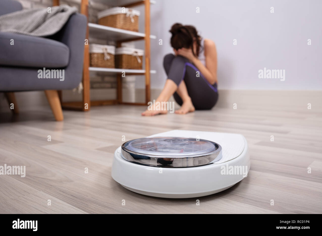 Close-up Of Weighing Scale In Front Of Sad Woman Sitting On Hardwood Floor Stock Photo