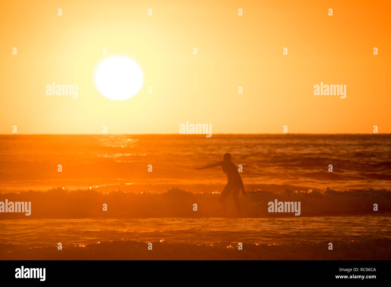A photo of a silhouette of a woman surfing on the background of a beautiful golden sunset. Jaco Beach, Costa Rica. Person is unrecognizable. Stock Photo