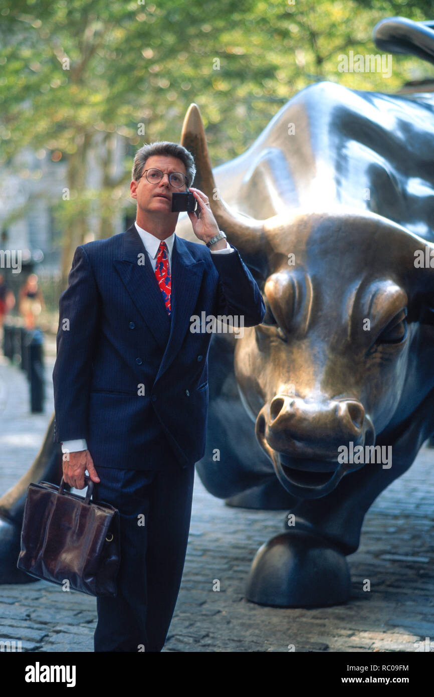 1990s businessman is on his mobile phone in front of the charging bull statue, Financial District, New York City, USA Stock Photo