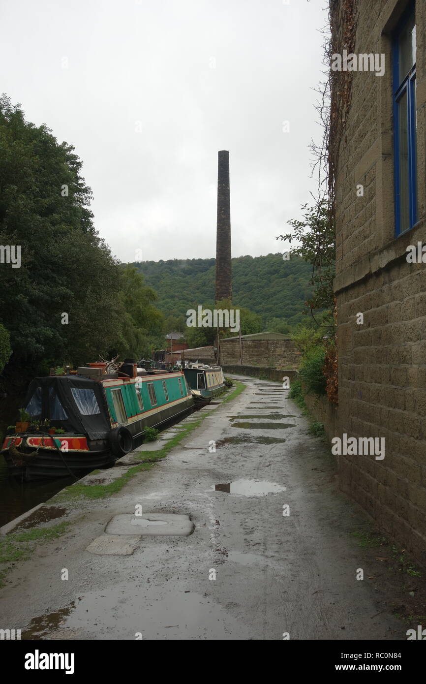 Canalside location in Hebden Bridge with traditional narrowboats on the Rochdale Canal and an old stone mill chimney in the distance. Stock Photo