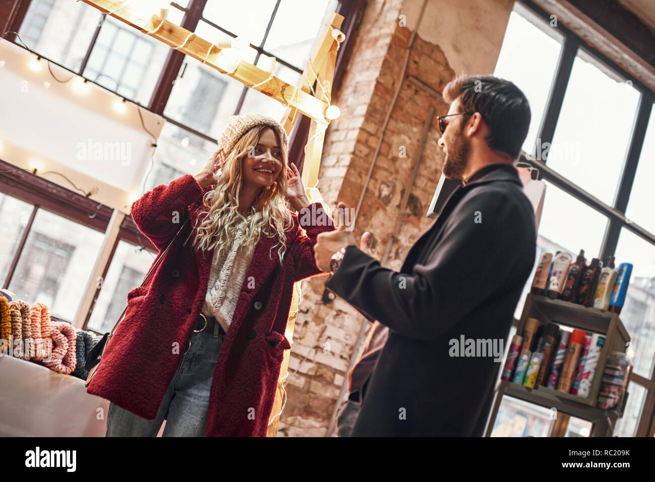 Good looking in this cap. Happy young joyful couple choosing caps at small street market. Autumn season, blond haired woman with her boyfriend are in  Stock Photo