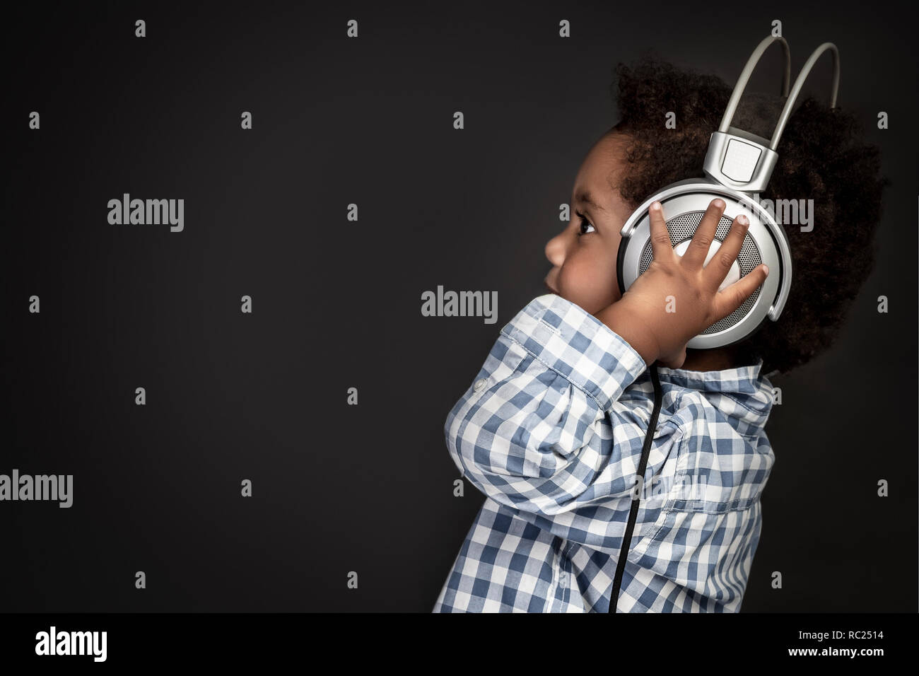 Profile portrait of a little curly African-American baby boy wearing big headphones and listening favourite music over black background, sweet child h Stock Photo