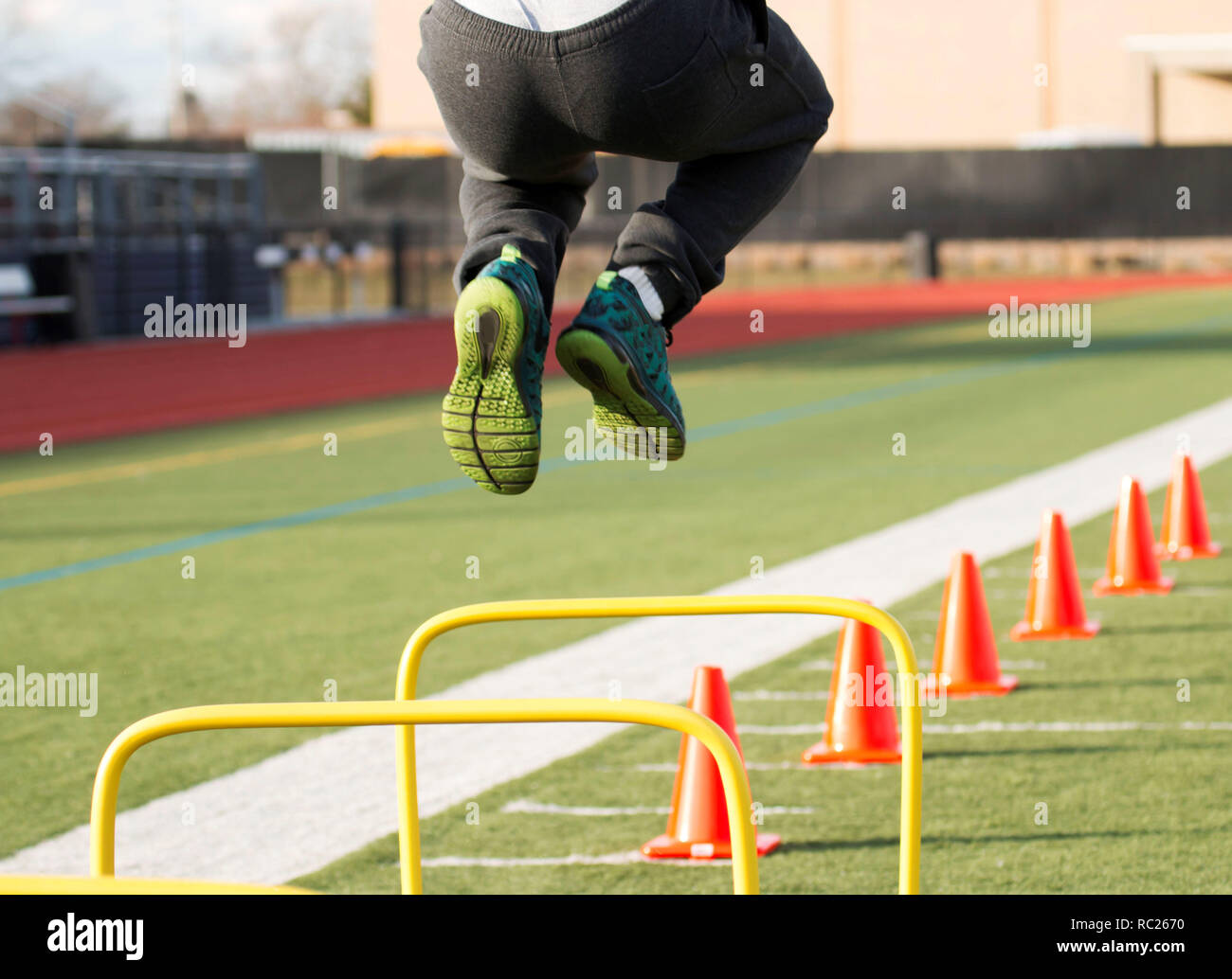 A track and field athlete jumps over yellow hurdles before he runs over orange cones, all on a green turf field Stock Photo
