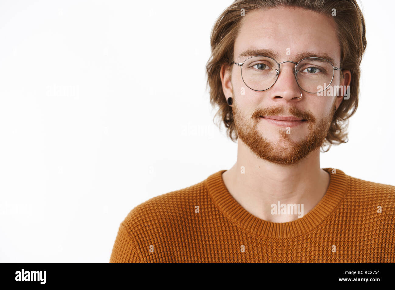 Close-up shot of charming happy and carefree bearded redhead guy with pierced nose in glasses and sweater standing on right side of copy space smiling looking hopefully at camera over gray wall Stock Photo