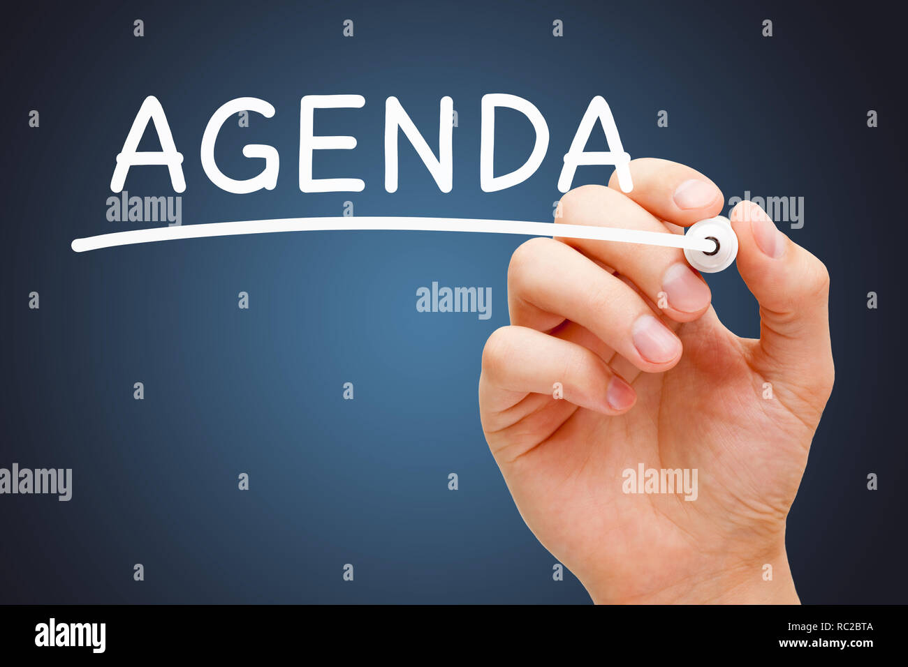 Hand writing the word Agenda with white marker on transparent wipe board over dark blue background. Stock Photo