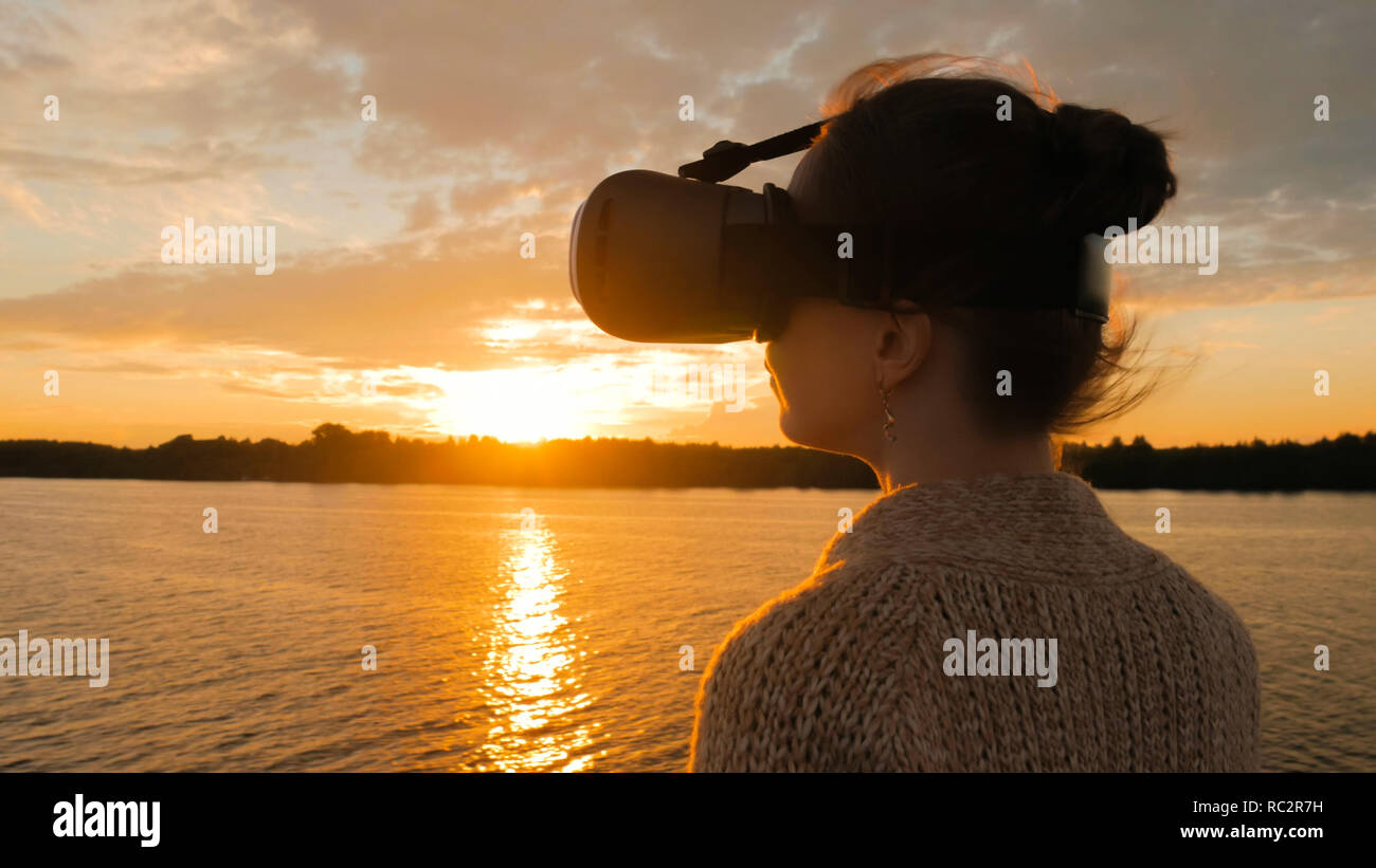 Woman using virtual reality headset on deck of cruise ship at sunset. Sunset light, golden hour, sun lens flares. Relax, future and technology concept Stock Photo