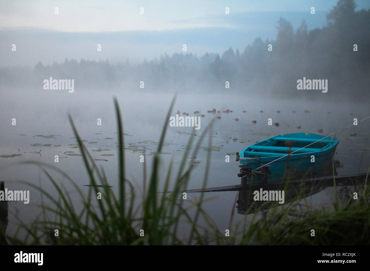 Lonely boat on a peaceful lake. Foggy autumn sunrise twilight. Bright blue turquoise shallop quiet forest, swamp plants, smoke mist on still calm water. Russia Moscow area nature landscape Stock Photo