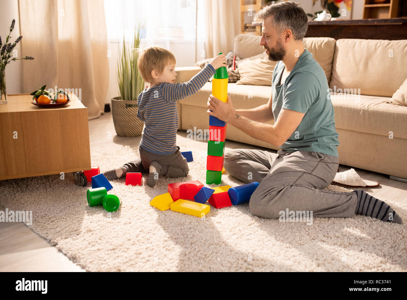 Curious cute little son in homewear sitting on carpet and putting cone on tower made of colorful cubes, bearded father and son playing with toy cubes Stock Photo