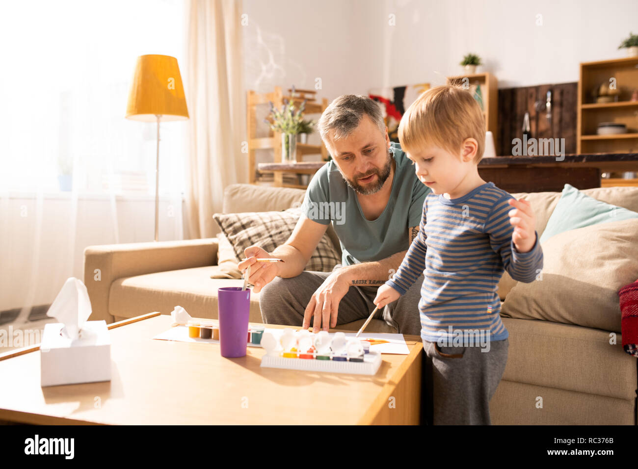 Serious handsome bearded single father sitting on sofa and teaching son to paint using acryl paints, curious boy paining picture with paintbrush Stock Photo