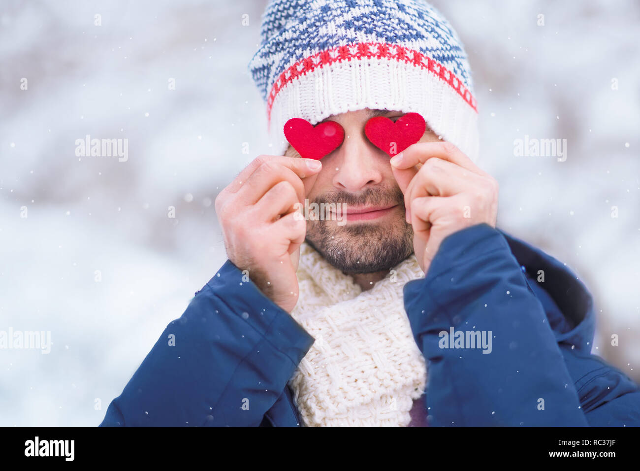 young caucasian man wearing blue jacket, hat and scarf holding two red hearts before his eyes outdoors for Valentines day Stock Photo