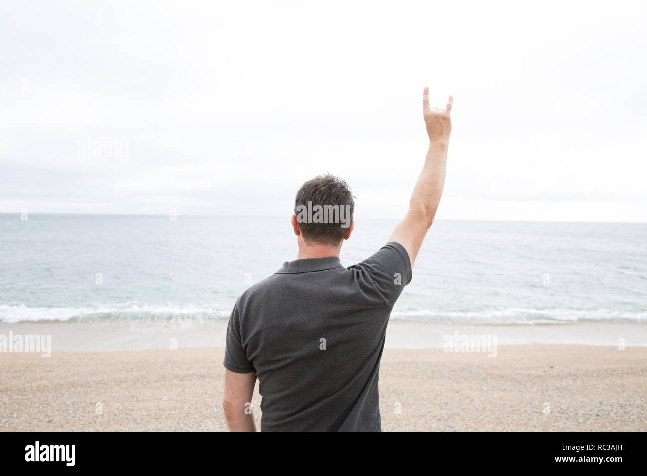 A rear view of a strong young man raising his arm in teh air and giving a devil horn gesture whilst facing the ocean in a symbol of hope and defiance  Stock Photo