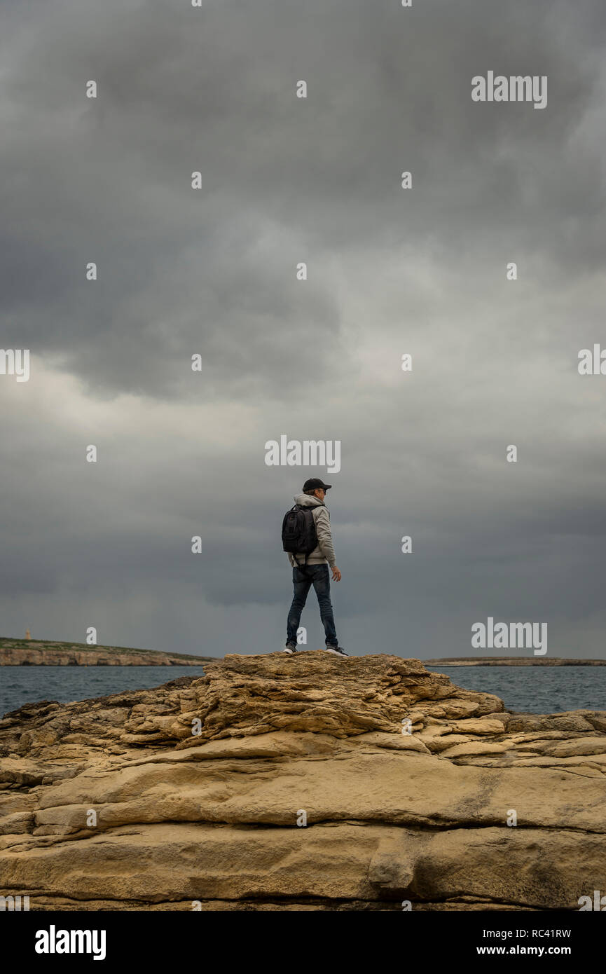 man with backpack standing on top of rocks looking out to sea, moody sky. Stock Photo