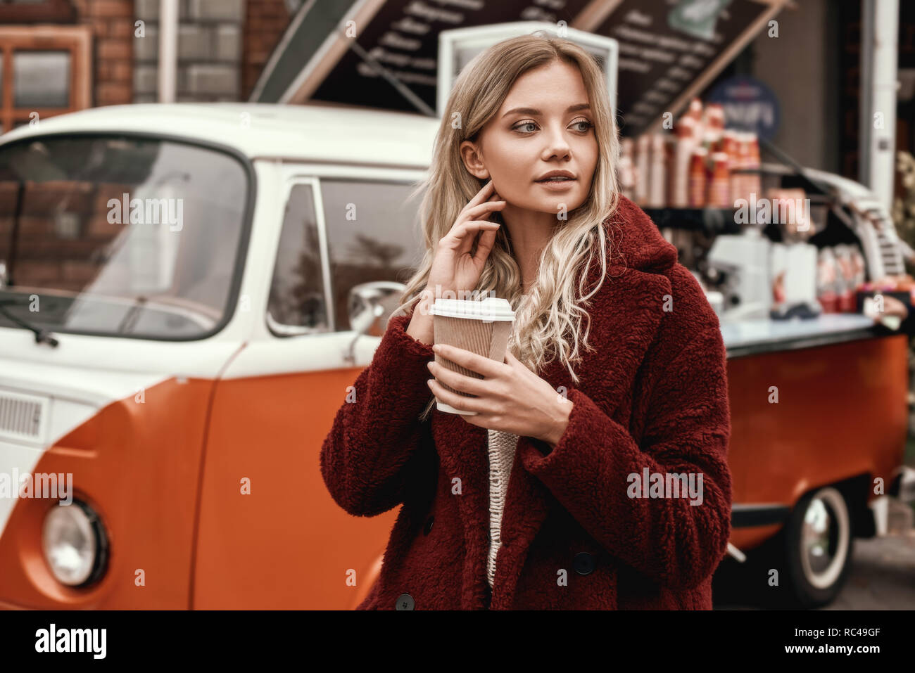 Drink coffee outdoors. Blonde beautiful white woman with long hair posing thougtfull on urban background and drinking coffee. Outdoor photo of lovable Stock Photo