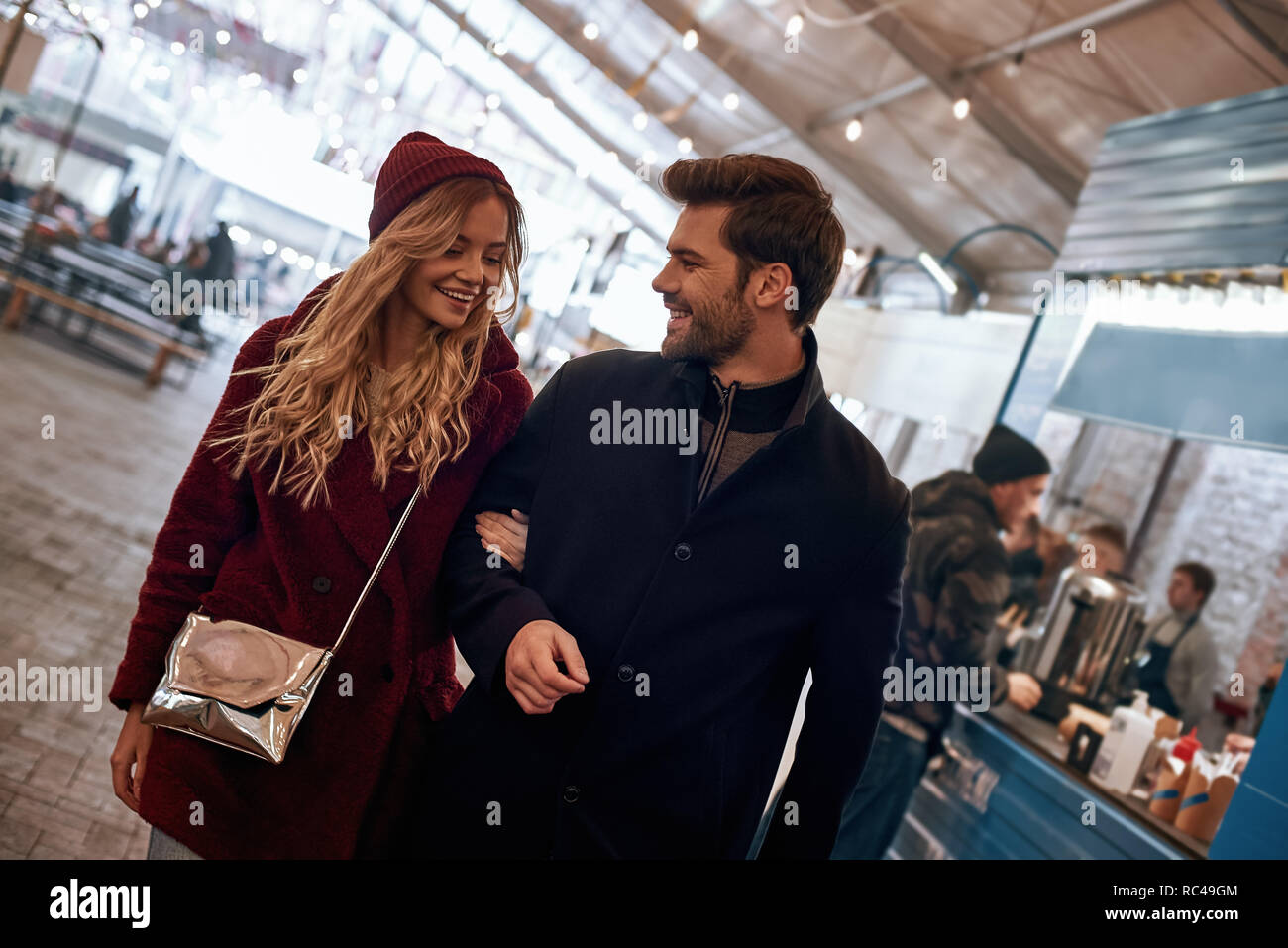 Stroll through the market together. Happy young joyful couple walking around street food market. Autumn season, blond haired woman wearing red cap, he Stock Photo
