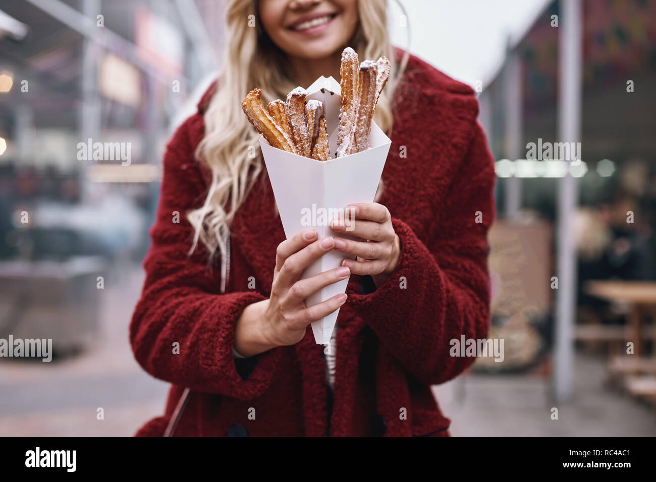 Street food culture. Young blonde woman in red cap and eco fur coat eatting churros at street fair. Stock Photo