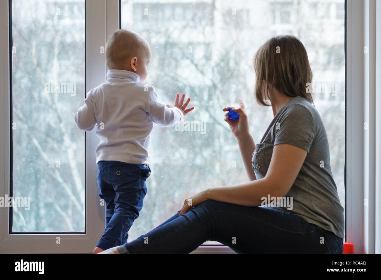 Mother and child are sitting on windowsill. They look out window. Winter day. People are unrecognizable. Stock Photo