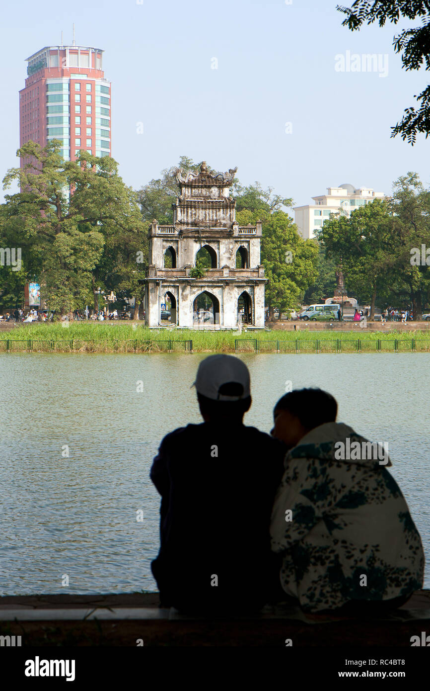 Defocused Vietnamese couple sitting by the Hoan Kiem Lake in Hanoi, Vietnam, on a sunny day. Focused on the old Turtle (Tortoise) Tower. Stock Photo